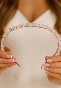 Przezroczysta opaska na głowę z cienką podstawą w kolorze różowego złota, z rzędem nieregularnie ukształtowanych kamieni na przedzie. Gładka faktura, minimalistyczny design.