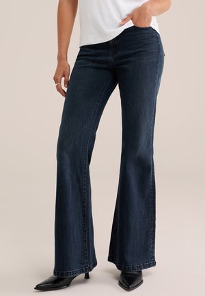 Femme portant un jean évasé bleu foncé, un haut blanc et des chaussures à talons noires à bout pointu, debout avec une main dans la poche.