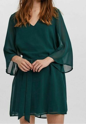 Femme portant une robe bleu sarcelle foncé à col en V avec des manches cloche en tissu transparent et une ceinture assortie nouée à la taille.