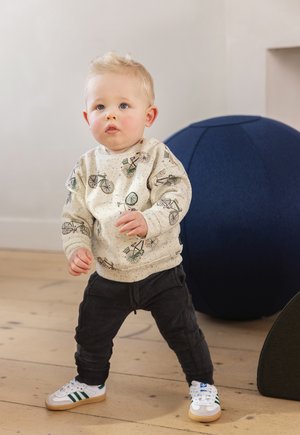 Hellbeige Sweatshirt mit Fahrrad-Print, dunkelgraue Jogginghose und weiße Sneakers mit grünen Akzenten. Stehend auf einem Holzboden.