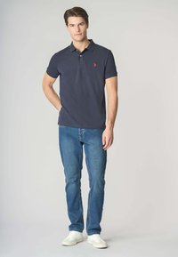 Polo shirt blu navy con un piccolo logo rosso, abbinato a jeans azzurri e sneakers bianche, caratterizzato da un colletto classico e maniche corte.