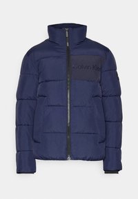 Marinblå pufferjacka med hög krage, dragkedjeslutning, texturerad yta och en framträdande Calvin Klein-logotyp på bröstet.