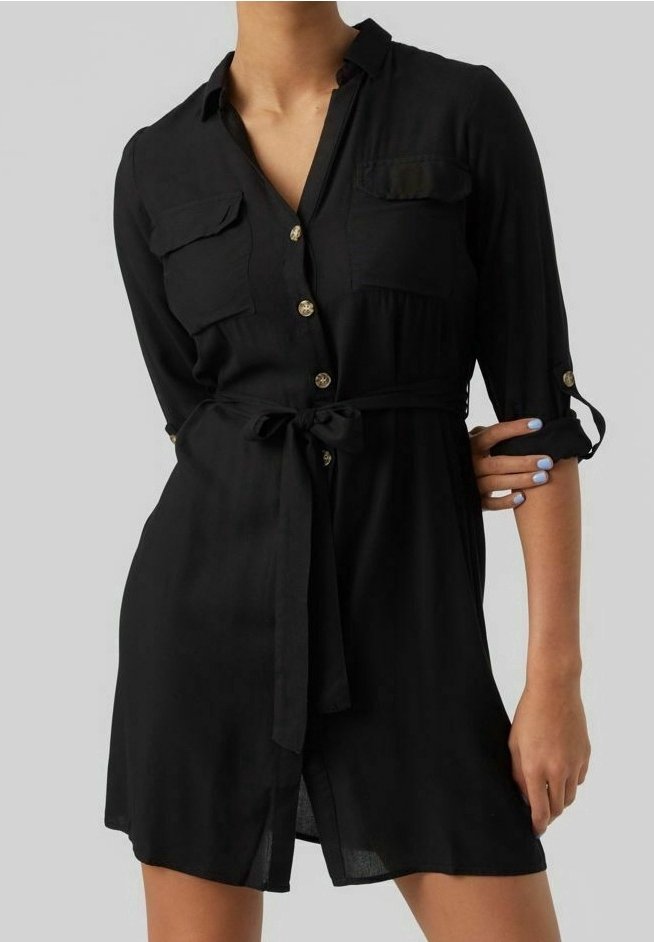 Femme portant une robe noire boutonnée avec des poches poitrine, une ceinture à la taille et des manches retroussées, debout devant un fond uni.
