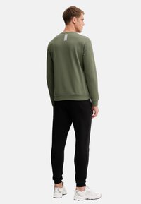 Felpa verde con scollo rotondo, caratterizzata da una striscia verticale bianca sul retro, abbinata a pantaloni jogger neri affusolati e sneakers bianche.