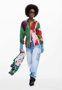 Buntes, gemustertes Langarmhemd in Grün, Pink und Braun, kombiniert mit hellblauen weiten Jeans und schwarzen Stiefeln mit Absatz; Modell hält einen Schal.