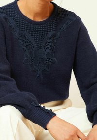 Pull court bleu marine avec broderie florale texturée autour de l'encolure et boutons décoratifs aux poignets. Tissu tricoté doux.