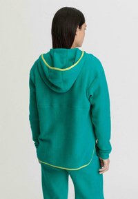 Felpa in pile color teal con rifiniture gialle, dotata di cappuccio e vestibilità comoda. La parte posteriore presenta un orlo curvo e due tasche laterali.
