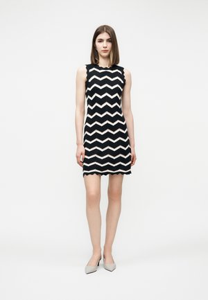 Kobieta stojąca, ubrana w czarną sukienkę bez rękawów z białymi wzorami w zygzaki i falistymi brzegami, w połączeniu z jasnoszarymi szpiczastymi szpilkami.