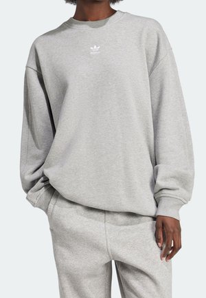 Personne portant un sweat-shirt Adidas gris clair et un pantalon de jogging assorti, les mains dans les poches, devant un fond uni.