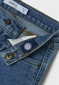 Jeans en denim bleu clair avec des coutures marron contrastées. Comprend une fermeture à bouton, une étiquette et un petit détail de poche.