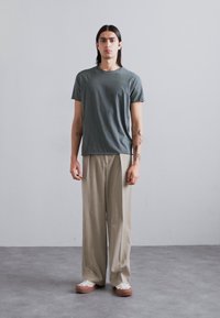 Grey cotton t-shirt with short sleeves, paired with beige wide-leg trousers and light footwear, standing against a neutral background.