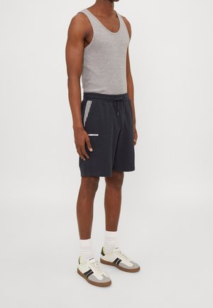 Mężczyzna w szarej koszulce bez rękawów, czarnych szortach na sznurki z paskiem bocznym, białych skarpetach i białych tenisówkach z czarnymi paskami.