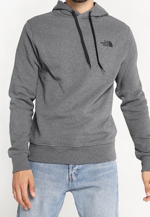 Mężczyzna w szarej bluzie z kapturem The North Face z czarnymi sznurkami i jasnoniebieskich jeansach, stojący na jednolitym tle.