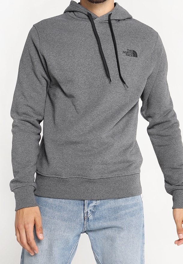 Homme portant un sweat à capuche gris The North Face avec des cordons noirs et un jean bleu clair, debout devant un fond uni.
