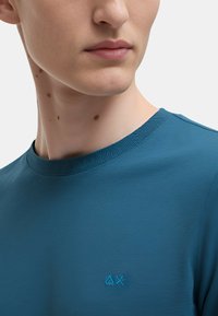 T-shirt blu a collo rotondo realizzato in tessuto morbido. I dettagli notevoli includono un logo sottile in blu chiaro sul petto e un collo a costine.