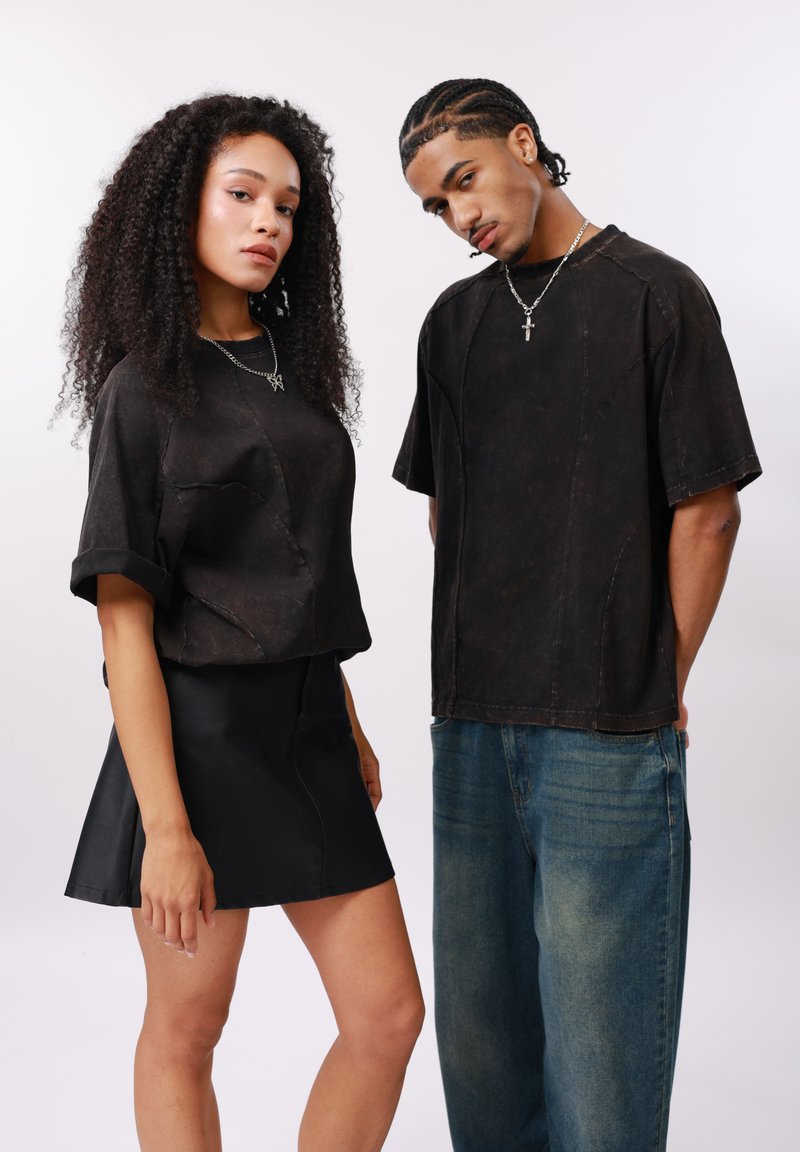 Νέα γυναίκα με σγουρά μαλλιά, φορεί μια μαύρη φούστα και μια φαρδιά μαύρη πουκαμίσα, στέκεται δίπλα σε νεαρό άνδρα με κομμένα κοτσίδα μαλλιά, που φοράει μαύρο πουκάμισο και μπλε τζιν.