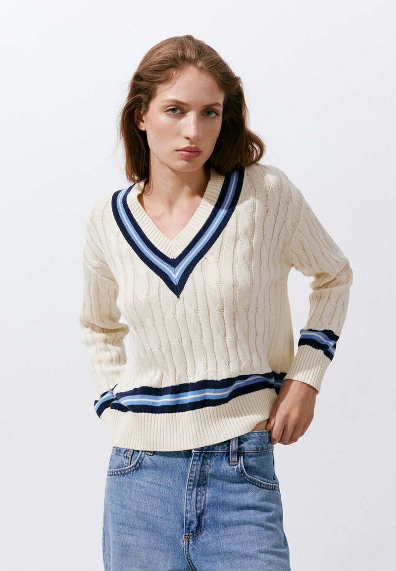 Jeune femme portant un pull en maille crème avec des rayures bleues sur le col en V, les poignets et l'ourlet, associé à un jean bleu, debout sur un fond blanc.