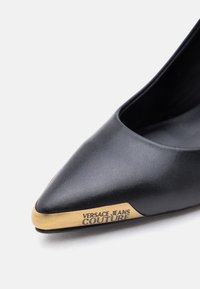 Μαύρο δερμάτινο παπούτσι μυτερό με χρυσό μεταλλικό διακοσμητικό στην άκρη με χαραγμένο το λογότυπο "Versace Jeans Couture".