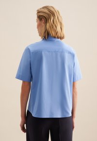 Koszula z krótkim rękawem w jasnoniebieskim kolorze, z kołnierzykiem, gładką fakturą i prostym dołem. Tył ma środkowy szew.