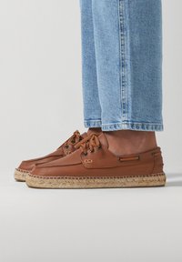 Brown leather boat shoes with matching laces and black eyelets. Espadrille-style jute sole. Worn with light blue cuffed jeans.