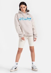 Beżowa bluza z kapturem z niebieskim grafiką, luźny krój, kieszeń kangurza. Dobrana z kremowymi szortami w prążki i szarymi trampkami.