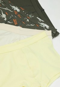 Drei Paar Jungs-Boxershorts in Gelb, beigeblümtem Streifenmuster und Dunkelgrün mit Dinosaurierskelett-Druck, übereinander auf einer weißen Oberfläche angeordnet.