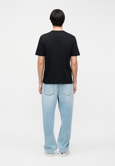 Άνδρας στέκεται με την πλάτη, φορώντας απλό μαύρο κοντομάνικο μπλουζάκι, ανοιχτόχρωμο μπλε τζιν χαλαρής εφαρμογής και λευκά αθλητικά παπούτσια σε λευκό φόντο.