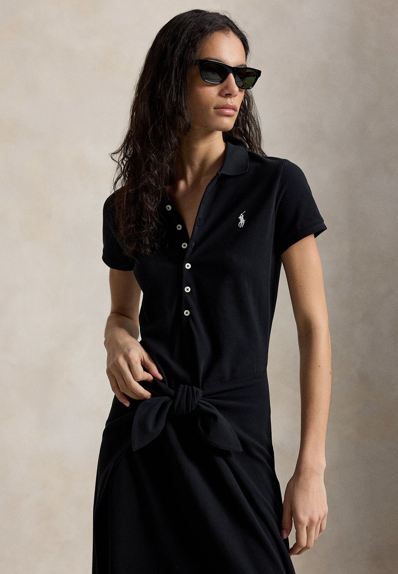 Žena v čiernych polo šatách s gombíkmi a logom, previazanými v páse, a čiernych slnečných okuliaroch, stojaca na neutrálom pozadí.