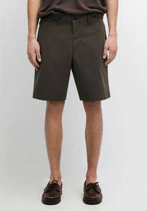 Uomo con pantaloncini marrone scuro fino al ginocchio con chiusura a bottone e zip, abbinati a mocassini casual marrone scuro, in piedi davanti a uno sfondo neutro.