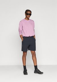 Ροζ ανοιχτόχρωμο μακρυμάνικο πουκάμισο με υφή λεπτομέρειας, σε συνδυασμό με σκούρα σορτς. Φοράει μαύρα αθλητικά παπούτσια και γυαλιά ηλίου, με τα χέρια στις τσέπες.