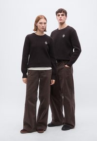 Pulls bruns en tricot côtelé, sous-maillots blancs et pantalons larges en velours côtelé brun ; logo géométrique notable sur les pulls, chaussures décontractées.