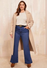 Palton bej cu un finisaj texturat, purtat peste un pulover deschis la culoare și blugi evazați până la talie de culoare albastră. Botine maro din suede completează aspectul.