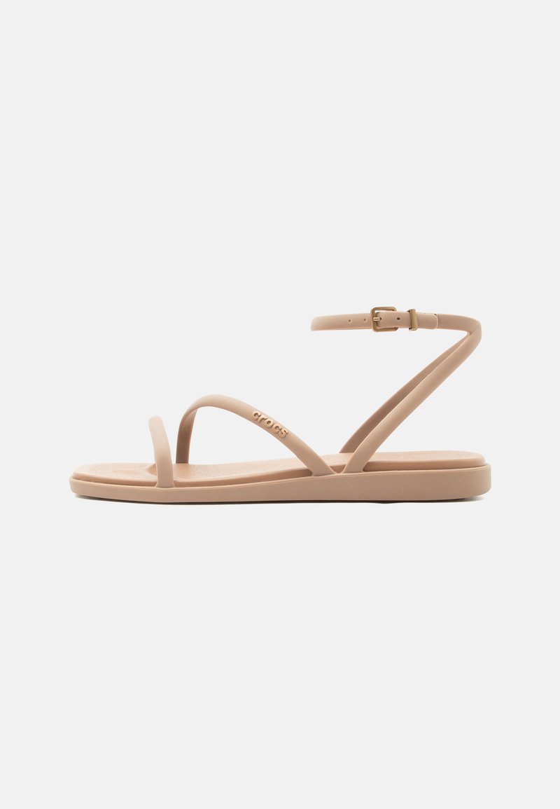 Beżowa płaska sandała z cienkimi paskami, klamrą przy kostce i pętlą na palec, zaprojektowana do codziennego noszenia.