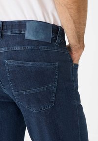Dark blue denim jeans with a slim fit, featuring a leather patch at the back, two back pockets, and subtle stitching details.