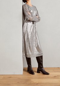 Robe en sequins argentés avec des manches longues, au design mi-genoux et un col rond. Associée à des bottes en cuir marron à talons hautes jusqu'aux genoux.