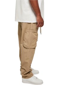 Beige cargo pants with a side pocket, made of cotton blend fabric. Worn with white athletic shoes. Casual design featuring a relaxed fit.