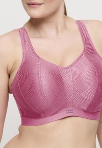 Nainen, jolla on yllään vaaleanpunainen kuvioitu tukevan rintaliivin, joissa on paksut hihnat ja brändätty elastinen nauha rinnan alla.