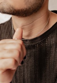 Un collier en chaîne argentée repose sur le cou, avec un design délicat et texturé. Un pull en tissu noir côtelé est partiellement visible, avec un col rond.