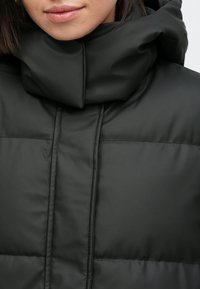 Čierna prešívaná bunda s vysokým, zloženým golierom. Hladká, lesklá textúra s viditeľným prešívaním a prešívanými panelmi. Minimalistický dizajn.