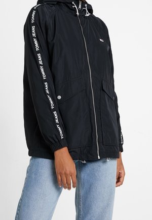 Musta kevyt takki hupulla, jossa on valkoinen "TOMMY JEANS" -brändäys hihoissa, etuvetoketju ja kaksi sivutaskua.