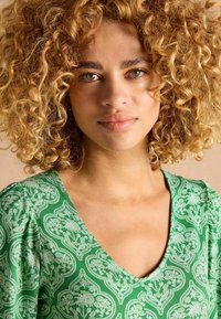 Zaļš v-veida krekls ar puffām piedurknēm, ar baltu ziedu paisley rakstu un gludu auduma tekstūru. Uzsver dekoltē līniju un plecus.