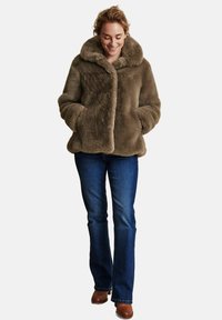 Brun fuskpälsjacka med bred krage, framknäppning och sidfickor, kombinerad med blå utsvängda jeans och bruna stövlar.