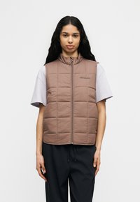 Γυναίκα με μακριά σκούρα μαλλιά που φοράει καφέ καπιτονέ γιλέκο πάνω από λευκό κοντομάνικο πουκάμισο και μαύρο παντελόνι, στέκεται σε απλό φόντο.