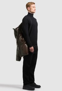 Homme vêtu entièrement de noir, debout de profil avec une doudoune marron foncé posée sur une épaule, sur un fond clair uni.