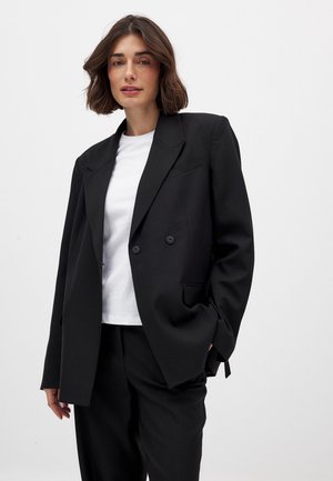 Musta kaksirivinen bleiseri, jossa on säädetty istuvuus, huippukaulus ja kaksi nappia. Vaatteen päällä valkoinen t-paita, yhdistetty rentoihin housuihin.