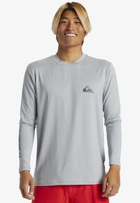 Dlhý rukáv, svetlošedý rash guard s okrúhlym výstrihom a priliehajúcim strihom. Obsahuje malé logo na hrudi a vzorované detaily na rukávoch.