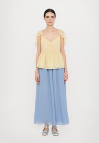 Gelbes Peplum-Top mit weichen, gerüschten Ärmeln, kombiniert mit hellblauen weiten Hosen. Silberne Schuhe vervollständigen das Outfit. Glatte Stofftextur.