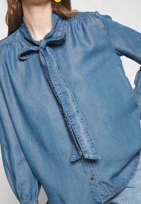 Femme portant une blouse en denim bleu clair avec un nœud au cou et des manches longues bouffantes, associée à un jean en denim bleu clair.