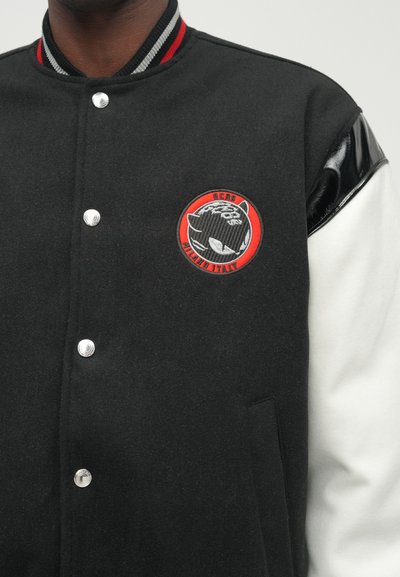 Black varsity jacket with a wool body, contrasting white sleeves, embroidered logo in red circle, snap buttons, and glossy shoulder accents.