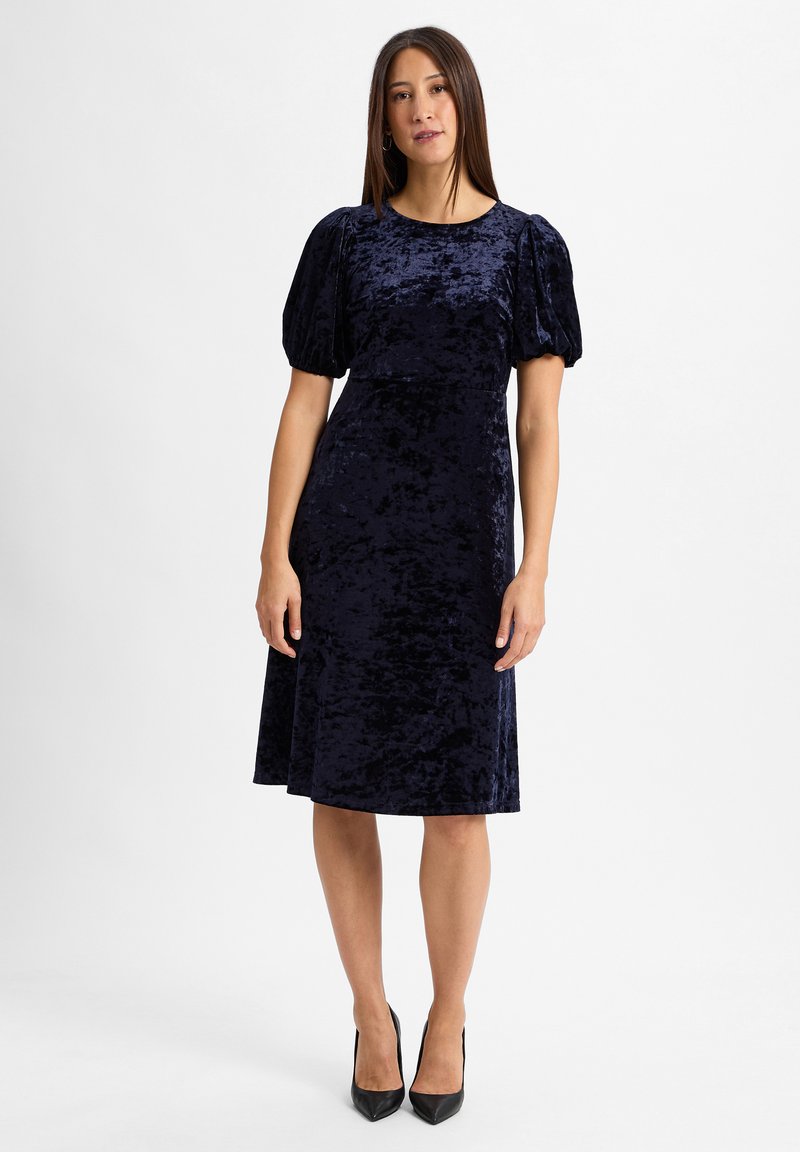 Dunkelblaues Samtkleid mit puffärmel und taillierter Passform, das bis zum Knie ausgestellt ist. Abgerundet mit eleganten schwarzen High Heels.